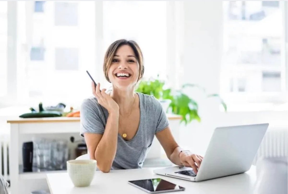 Smiling woman at desk with laptop, holding pencil, bright modern office with plants and window view