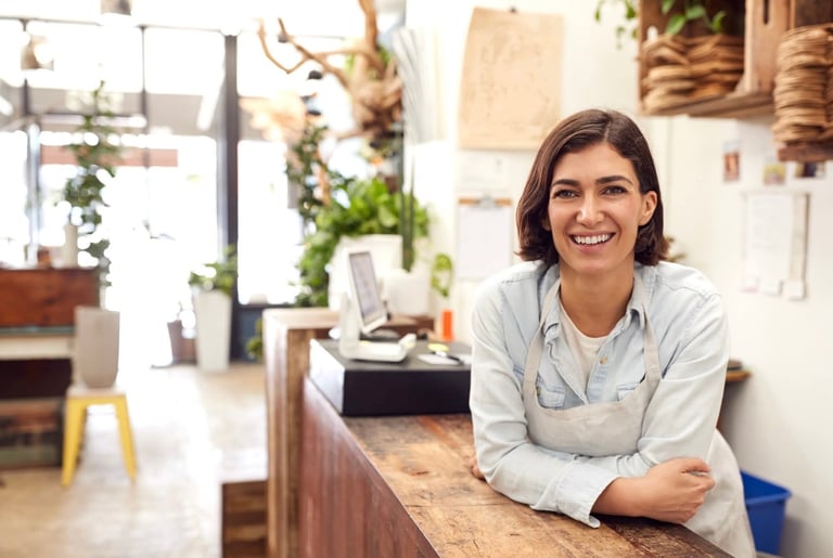 Smiling woman in apron leaning against wooden counter in a bright, plant-filled studio or shop space